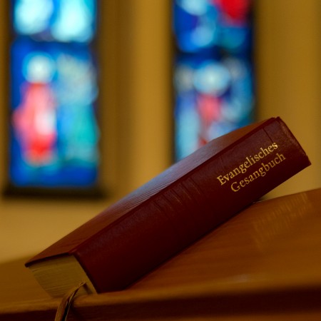 Evangelisches Gesangbuch auf einer Kirchenbank liegend. Im Hintergrund sind bunte Kirchenfenster zu sehen