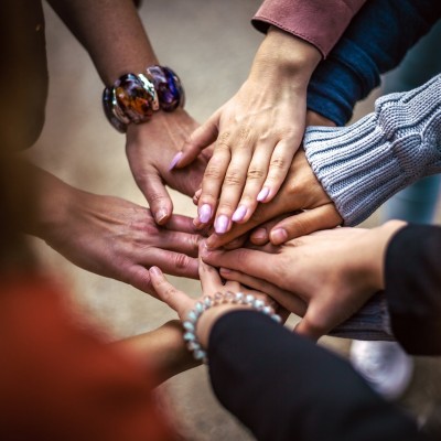 Mehrere Hände liegen übereinander in der Mitte, als Zeichen für Zusammenhalt und Teamgeist – verschiedene Armbänder und Ärmel sichtbar