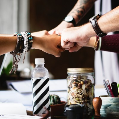 Mehrere Hände stoßen in der Mitte zusammen zu einer Faustgeste über einem Tisch mit Wasserflasche, Glasbehälter mit Nüssen, kleiner Pflanze und Büroartikeln – Symbol für Teamwork und Zusammenarbeit